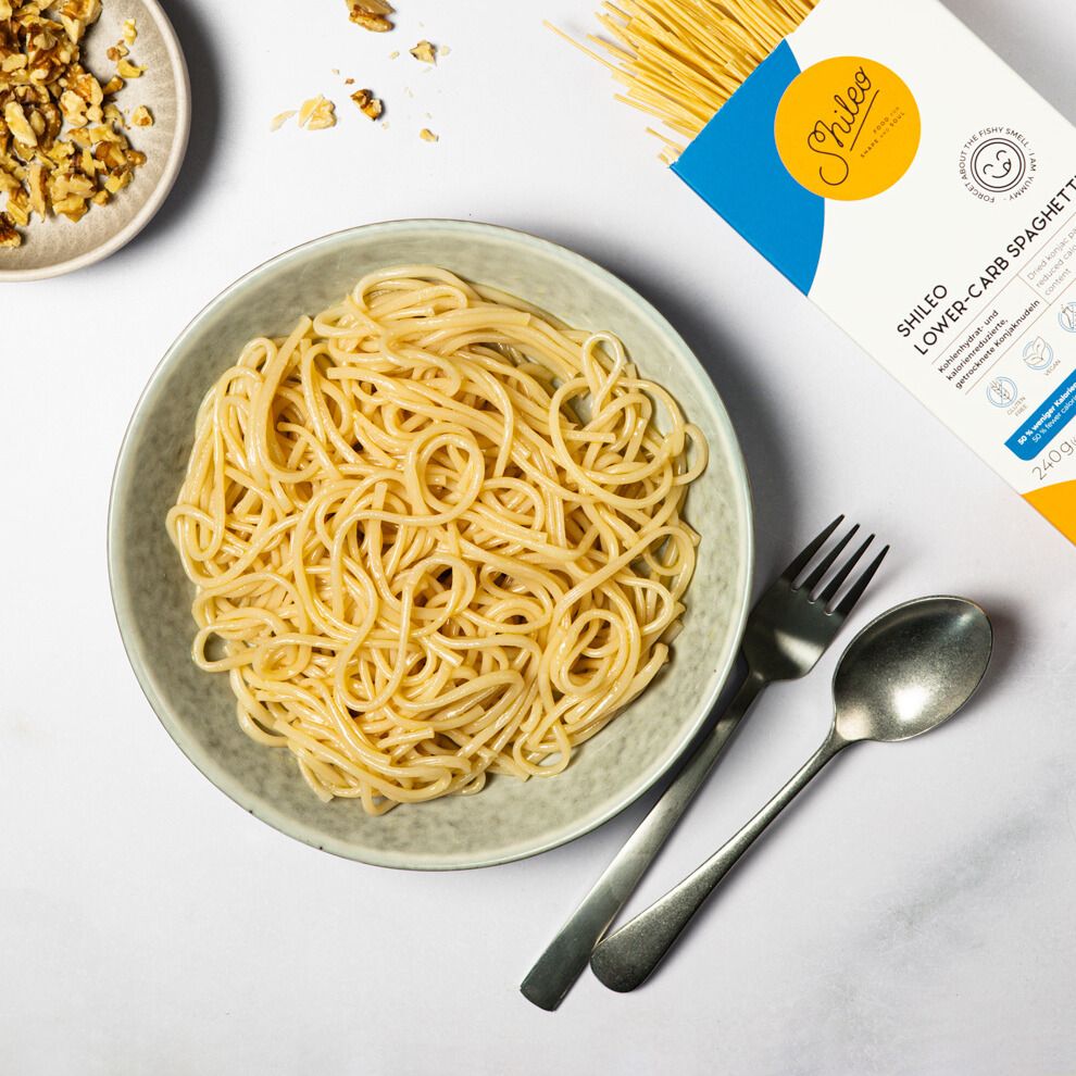 Teller mit gekochten Spaghetti. Daneben eine Packung SHILEO Lower-Carb Spaghetti und Besteck. Walnüsse in einer Schale.