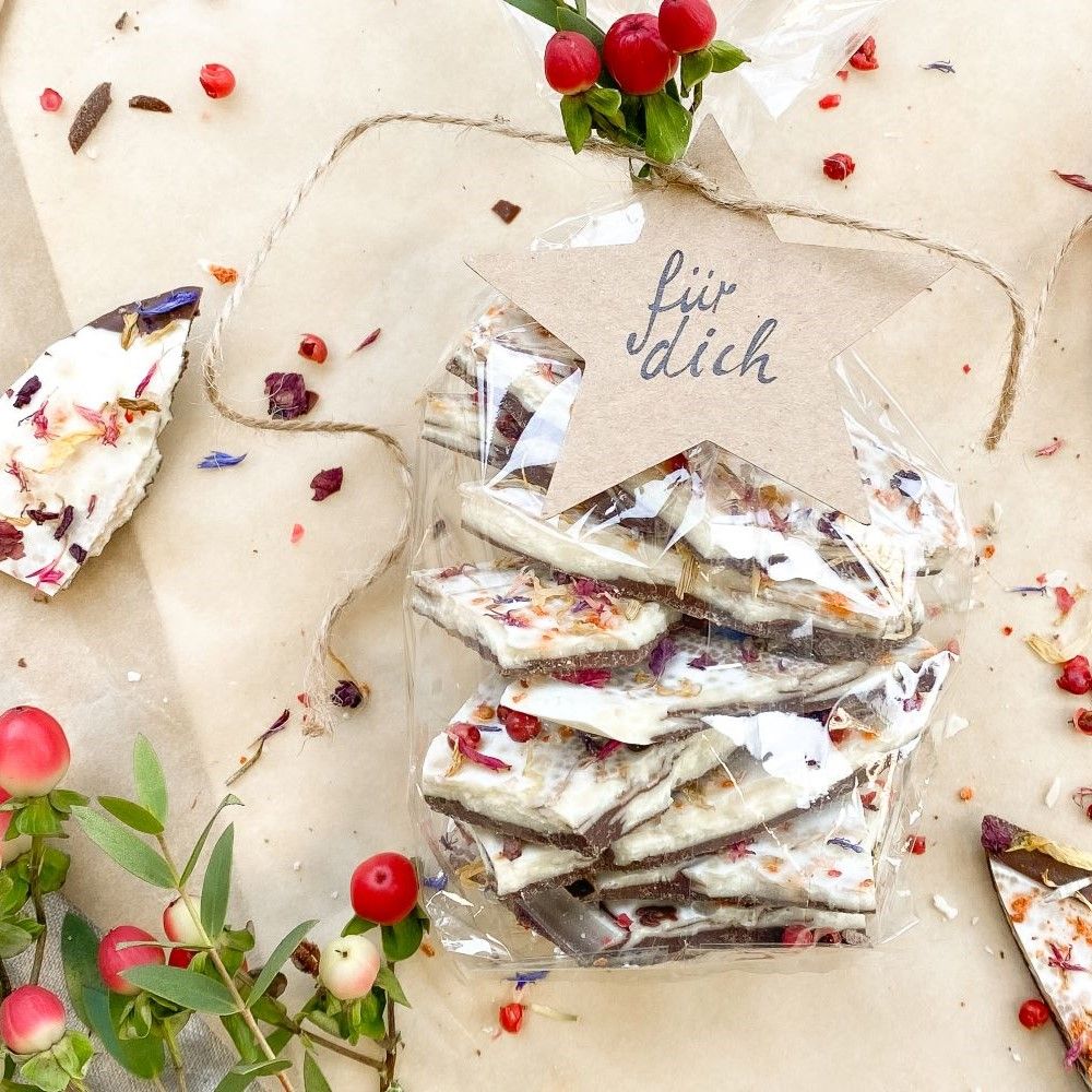 Transparente Tüte mit Bruchschokolade und Blüten. Sternförmiges Etikett mit Schriftzug. Dekoration mit Beeren.