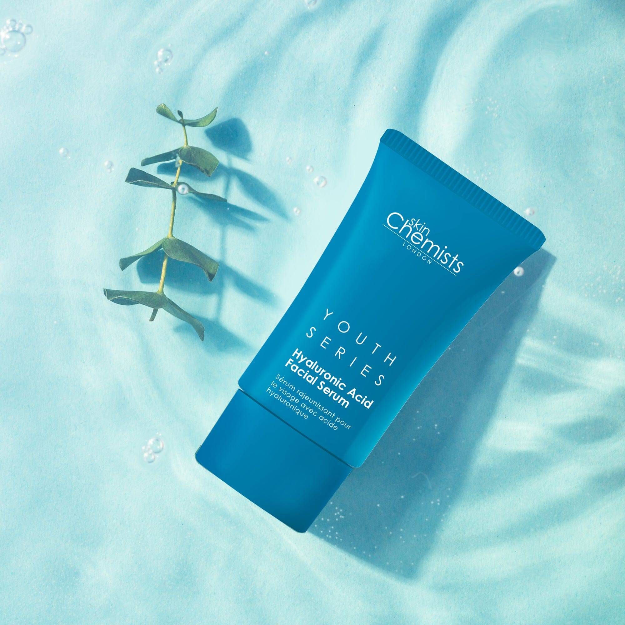 Blaues Tube mit "skinChemists"-Logo und "Youth Series Hyaluronic Acid Facial Serum"-Text, platziert auf einer Oberfläche mit Wasser und Pflanze.