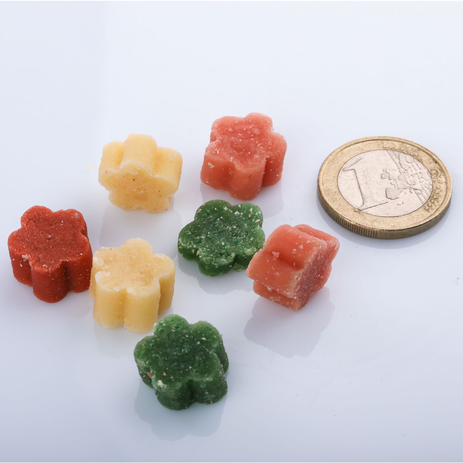 Mehrfarbige, blütenförmige Hundesnacks. Sieben Stück liegen neben einer 1-Euro-Münze. Farben: rot, gelb und grün. Nahaufnahme auf weißem Hintergrund.