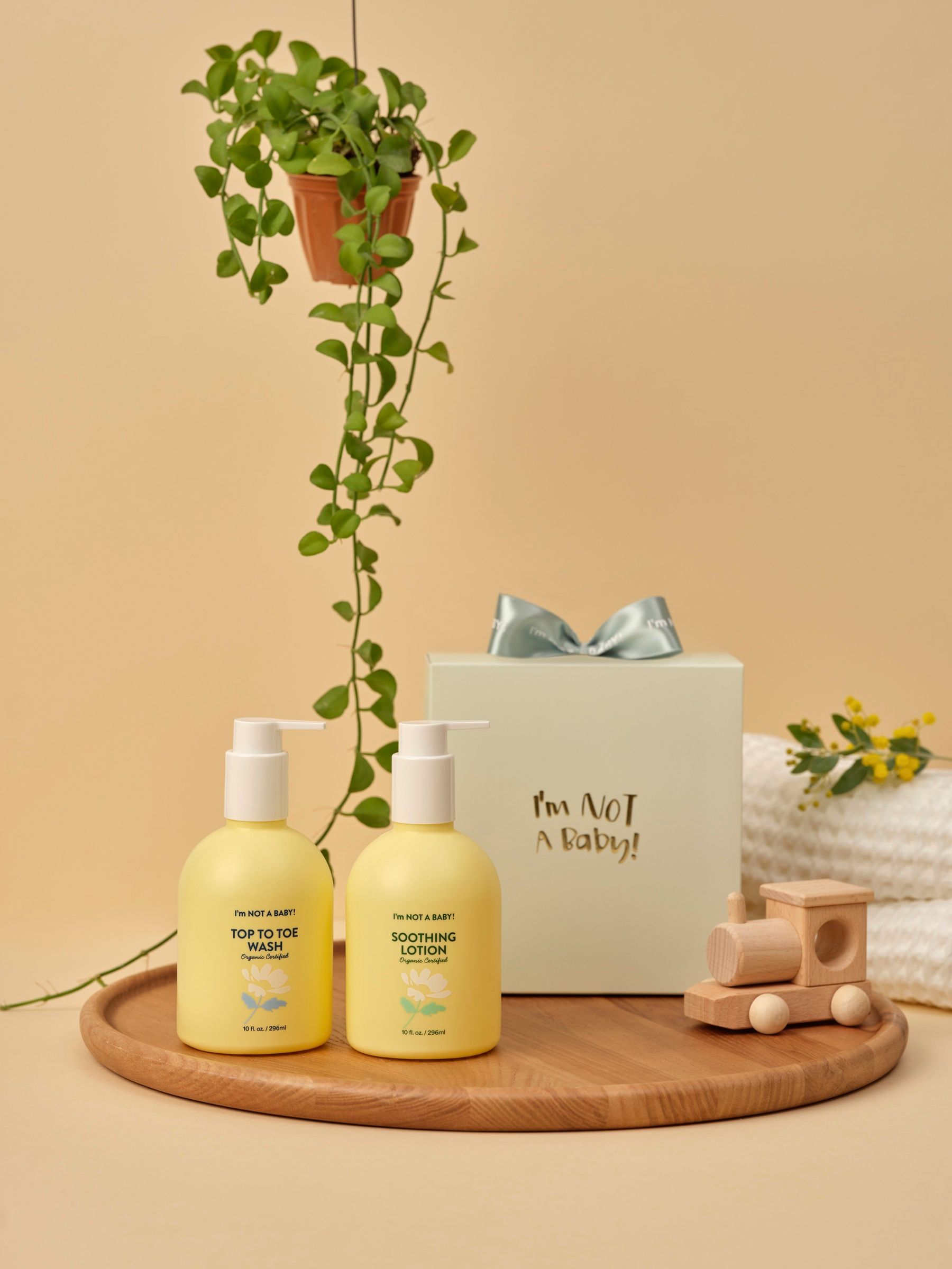 Zwei gelbe Flaschen und eine Geschenkbox mit Schleife auf einem Holztablett. Aufschrift: I'm NOT A BABY. Daneben ein Holzzug und Handtücher.