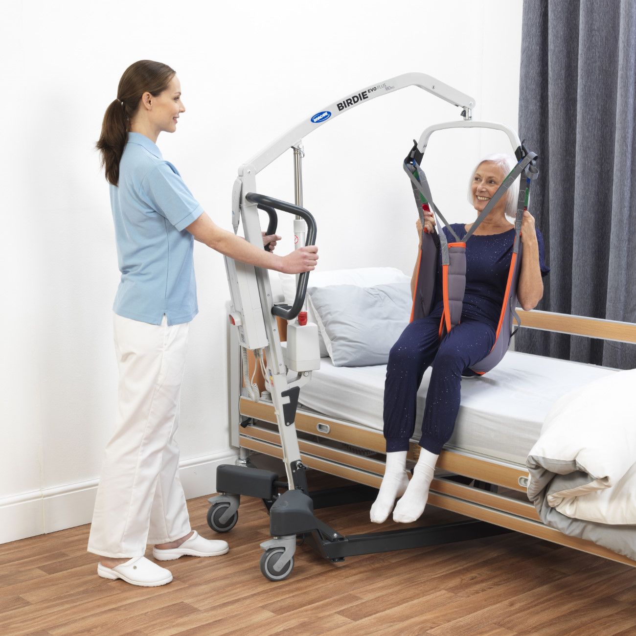 Patientin sitzt im Gurt des Lifters. Pflegekraft bedient den Lift. Invacare Birdie Evo Plus.