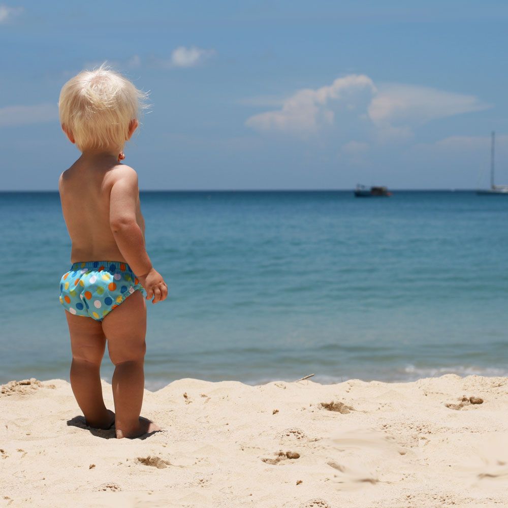 Baby steht am Strand, trägt eine Schwimmwindel mit blauem Muster. Blick auf Meer und Himmel.