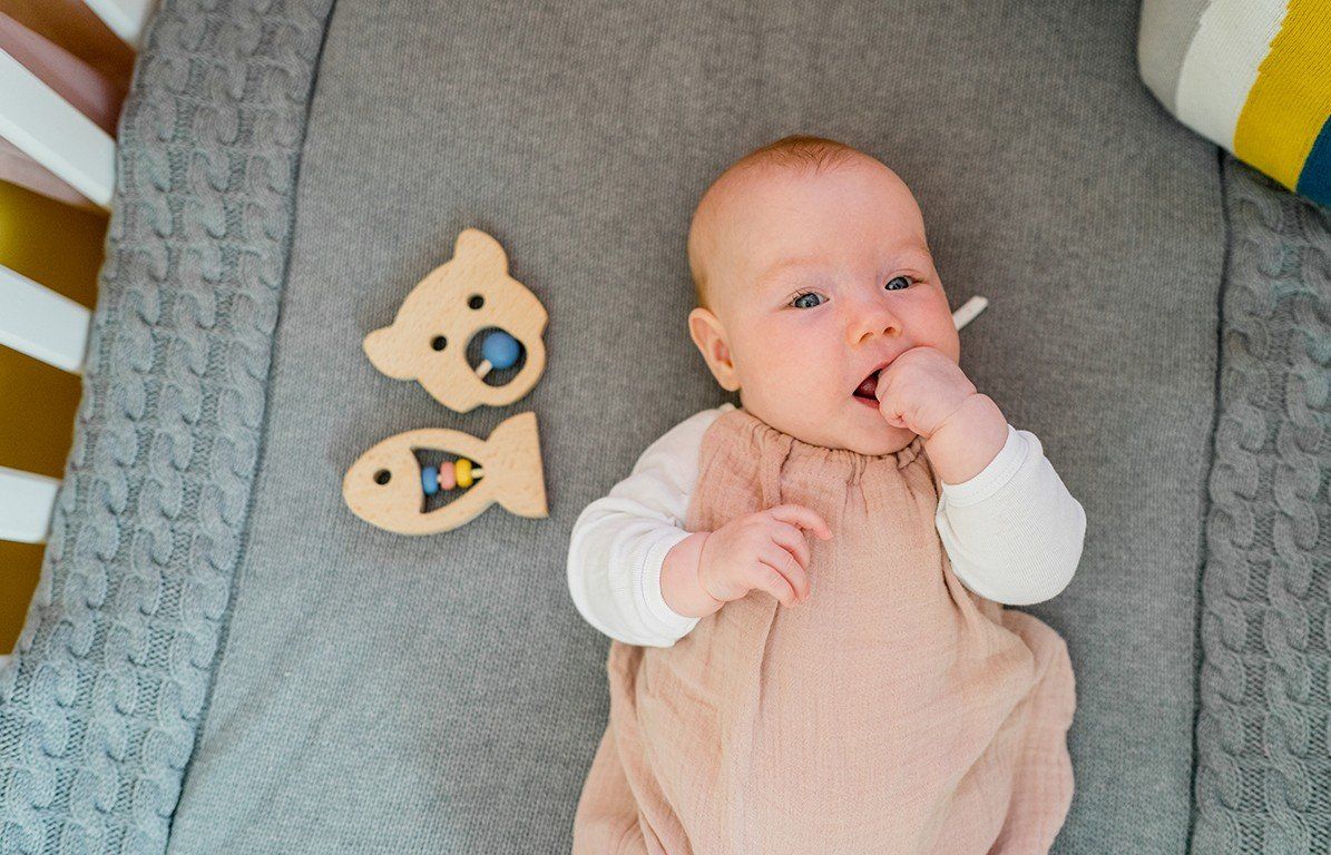 Baby liegt auf einer grauen Decke. Neben ihm liegen zwei Holz-Beißringe: Bär und Fisch. Baby steckt Finger in den Mund.