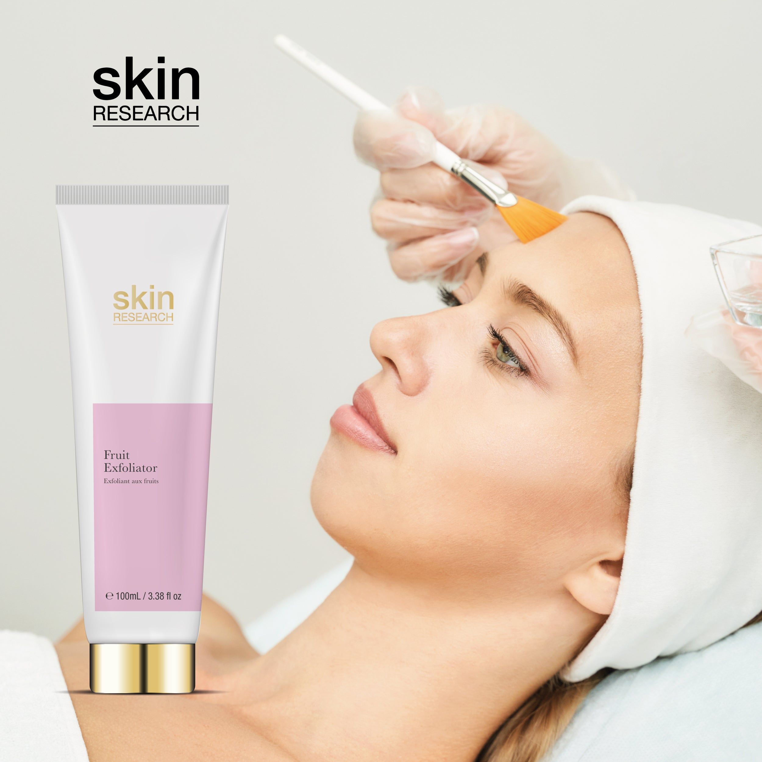 Weiße Tube mit rosa Etikett und goldfarbenem Verschluss, neben einer Frau, die eine Gesichtsbehandlung erhält. Auf der Tube steht "skin research".