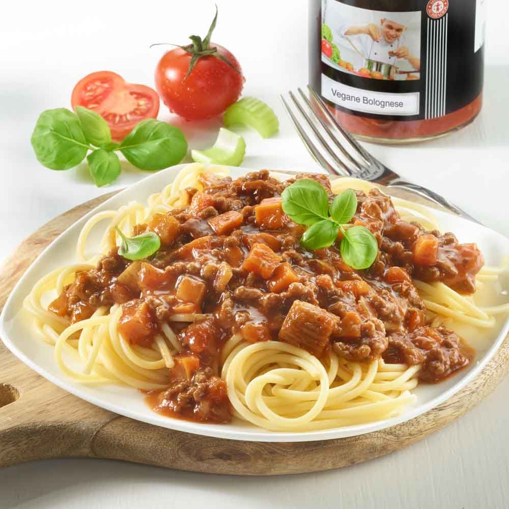 Teller Spaghetti mit Schrader Vegane Bolognese. Tomate, Basilikumblätter. Auf Holzbrett. Im Hintergrund Glas mit Etikett.