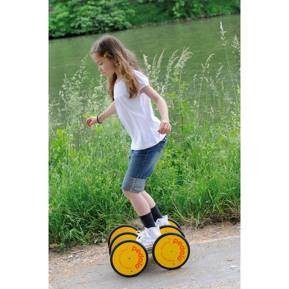 Mädchen auf einem Koordinationstrainer. Gelbe Räder mit "pedalo"-Logo. Sie trägt Jeansshorts und balanciert auf einem Weg.