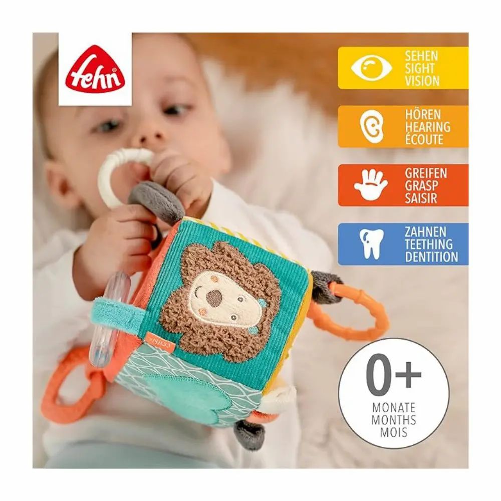 Baby hält Stoffwürfel mit Löwenmotiv. Marke 'Fehn' sichtbar. Hinweise zu Sehen, Hören, Greifen, Zähnen. Alter: 0+ Monate.
