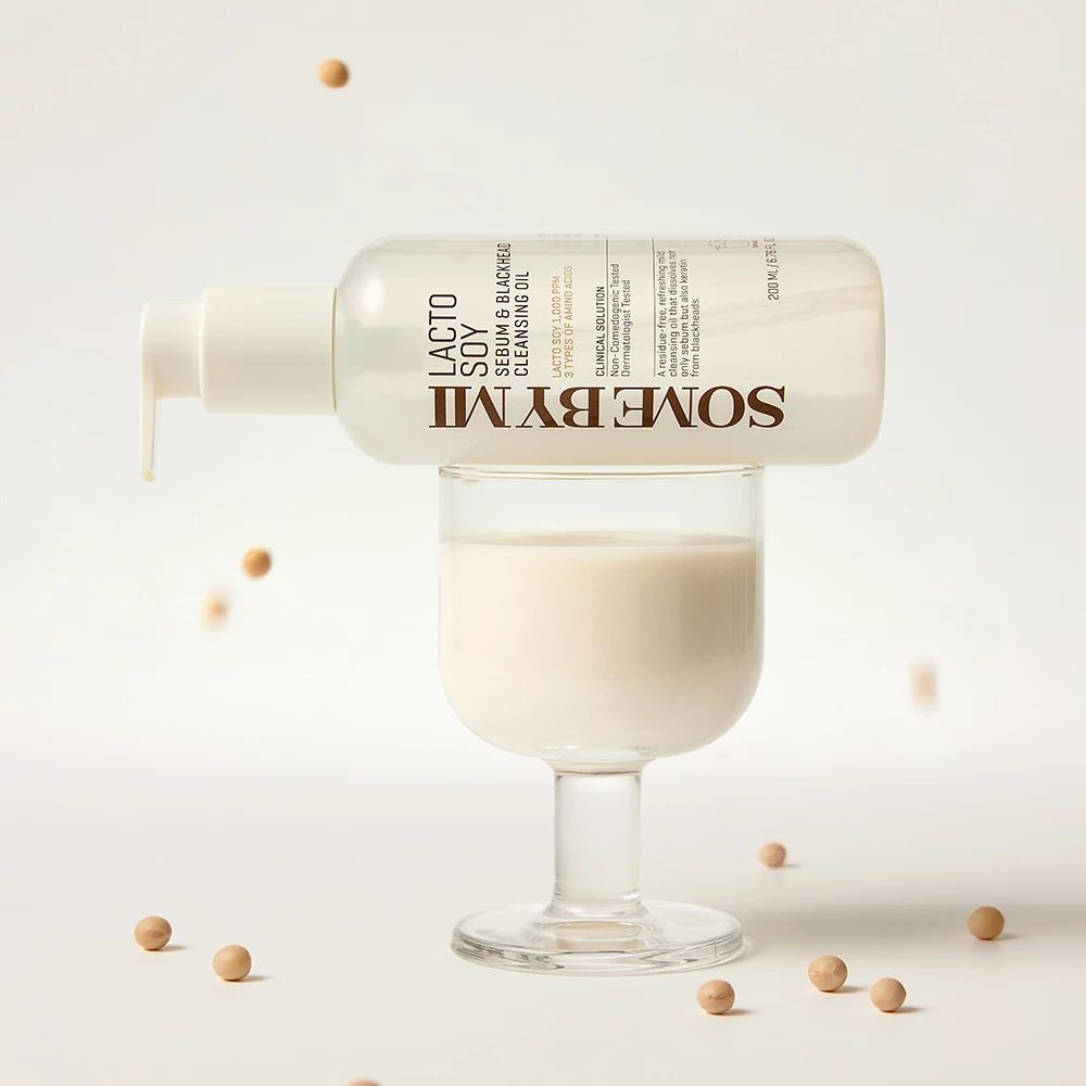 Klarflasche mit Pumpe, gefüllt mit Flüssigkeit, auf einem Glas. Aufschrift: SOMEBYMI Lacto Soy Sebum & Blackhead Cleansing Oil. Sojabohnen liegen herum.