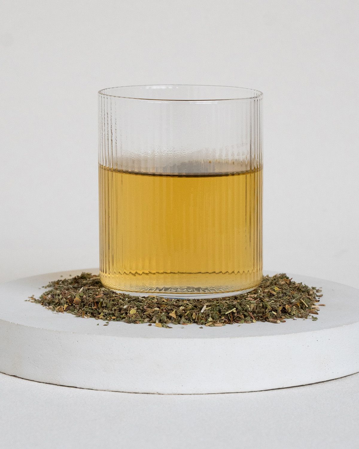 Glas mit Flüssigkeit und Kräutern. Geriffeltes Glas auf weißem Podest mit Kräutern. Flüssigkeit gelblich.