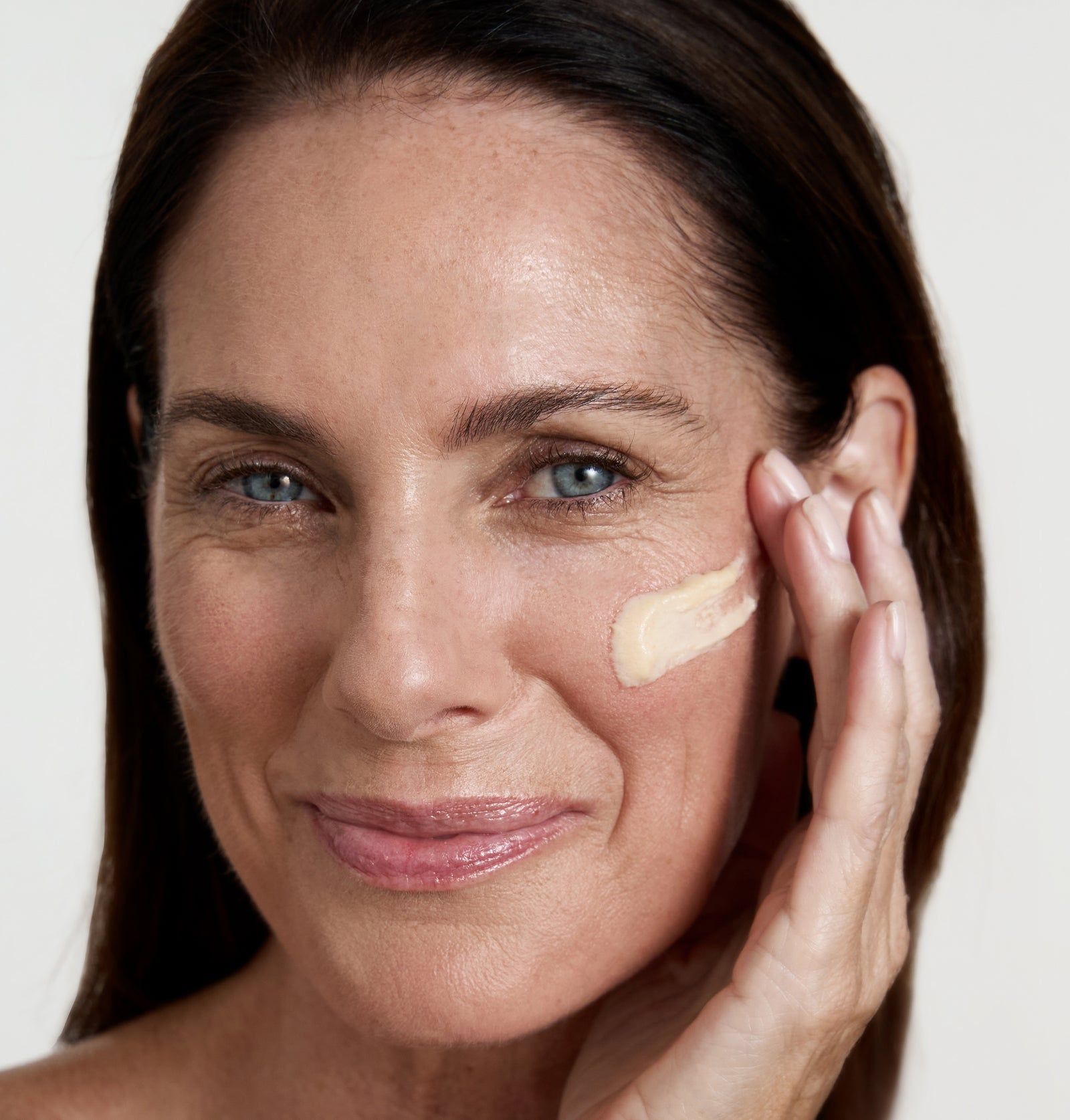 Frau mit Creme auf der Wange. Creme-Textur sichtbar. Helle Haut, dunkle Haare.