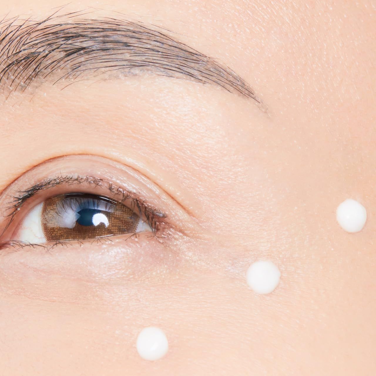 Nahaufnahme eines Auges mit drei weißen Creme-Punkten unter dem Auge. Hautfarbe, Augenbraue und Wimpern sichtbar.
