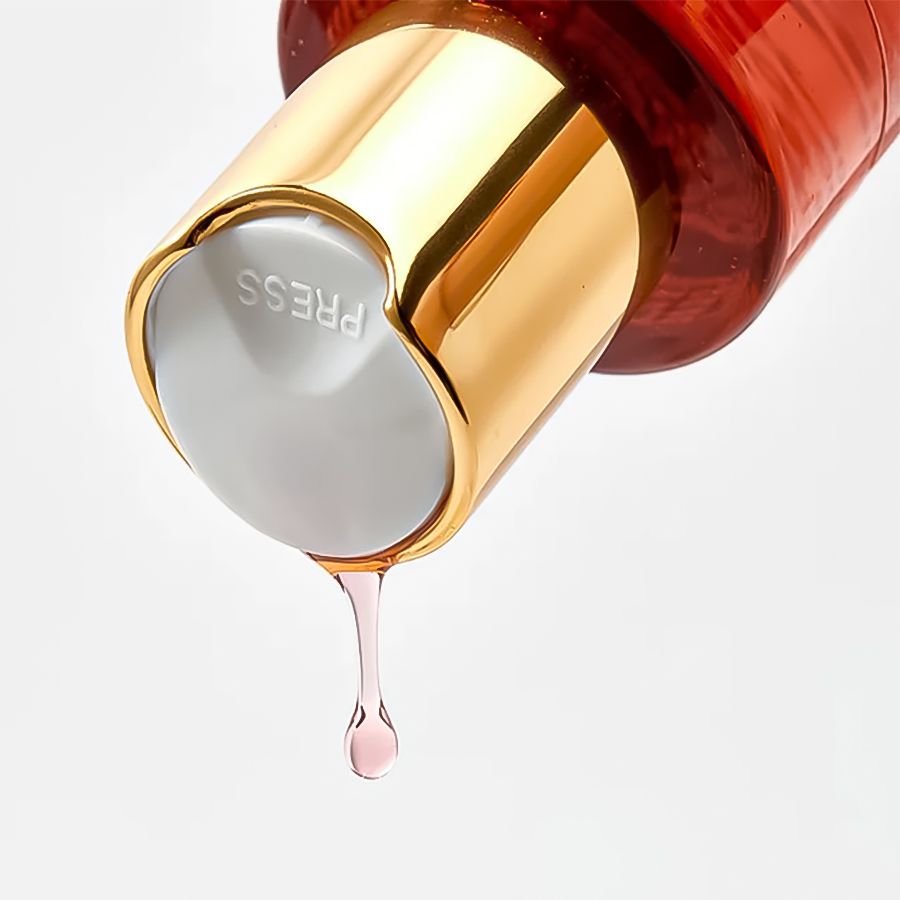 Goldfarbener Verschluss mit grauem Knopf. Aufschrift: PRESS. Tropfen rosa Flüssigkeit.