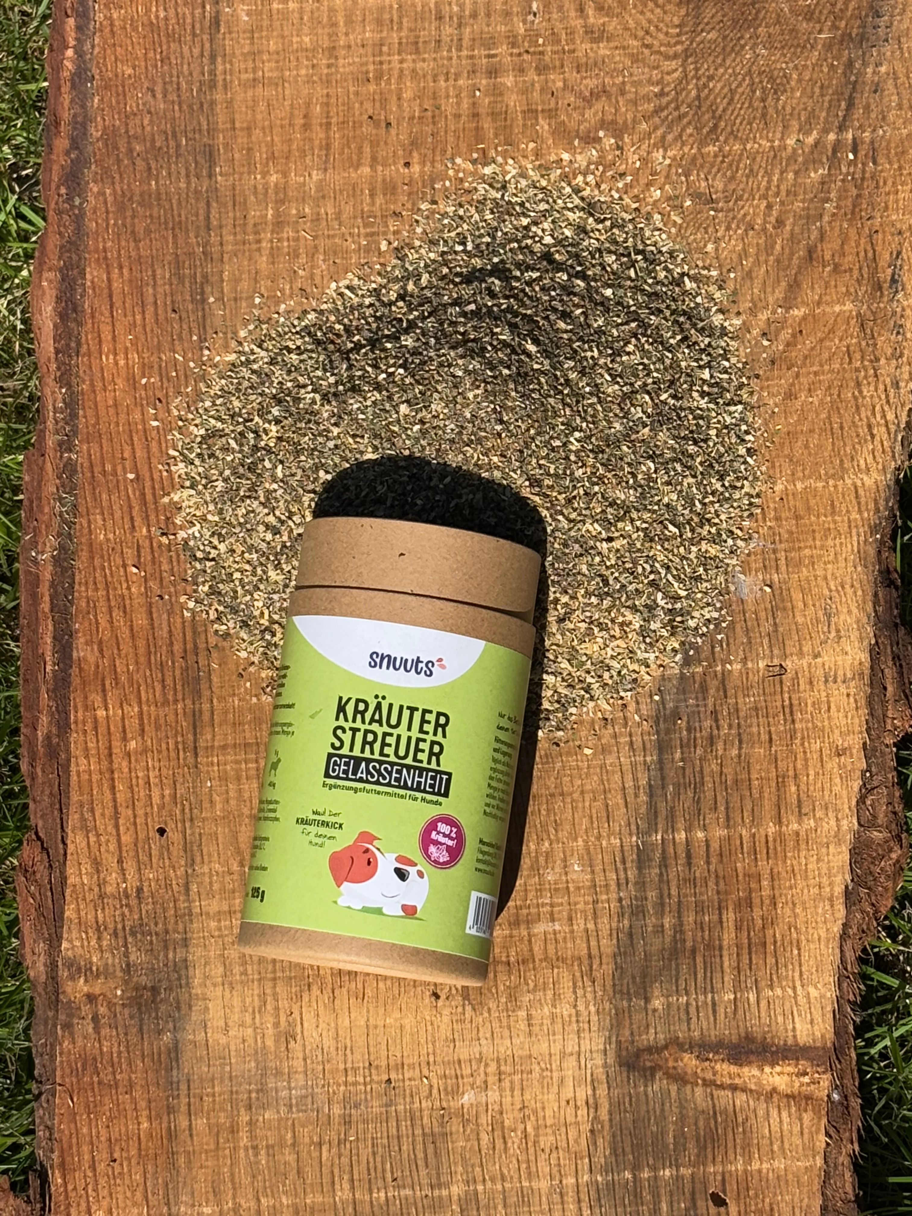 Kräuterstreuer-Dose und Kräuterhaufen auf Holz. Dose mit grünem Etikett, Aufschrift: snuuts Kräuterstreuer Gelassenheit. Brauner Deckel. Kräuterstreuer-Inhalt daneben.