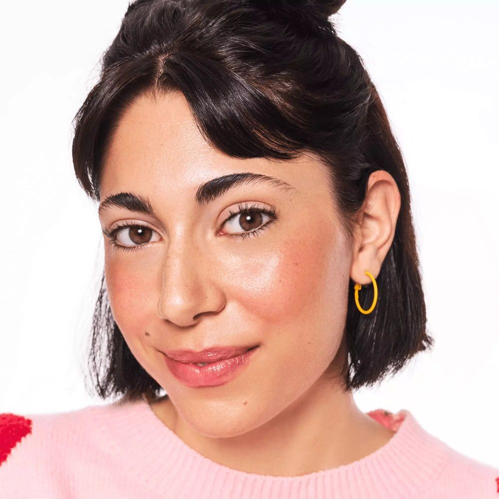 Porträt einer Frau mit gebräunter Haut. Sie trägt gelbe Ohrringe und rosa Pullover.