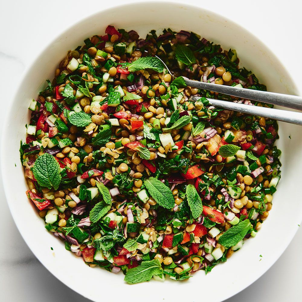 Salat mit Linsen, Tomaten, Gurken, Zwiebeln und Minze in einer weißen Schüssel. Zwei silberne Zangen liegen im Salat.