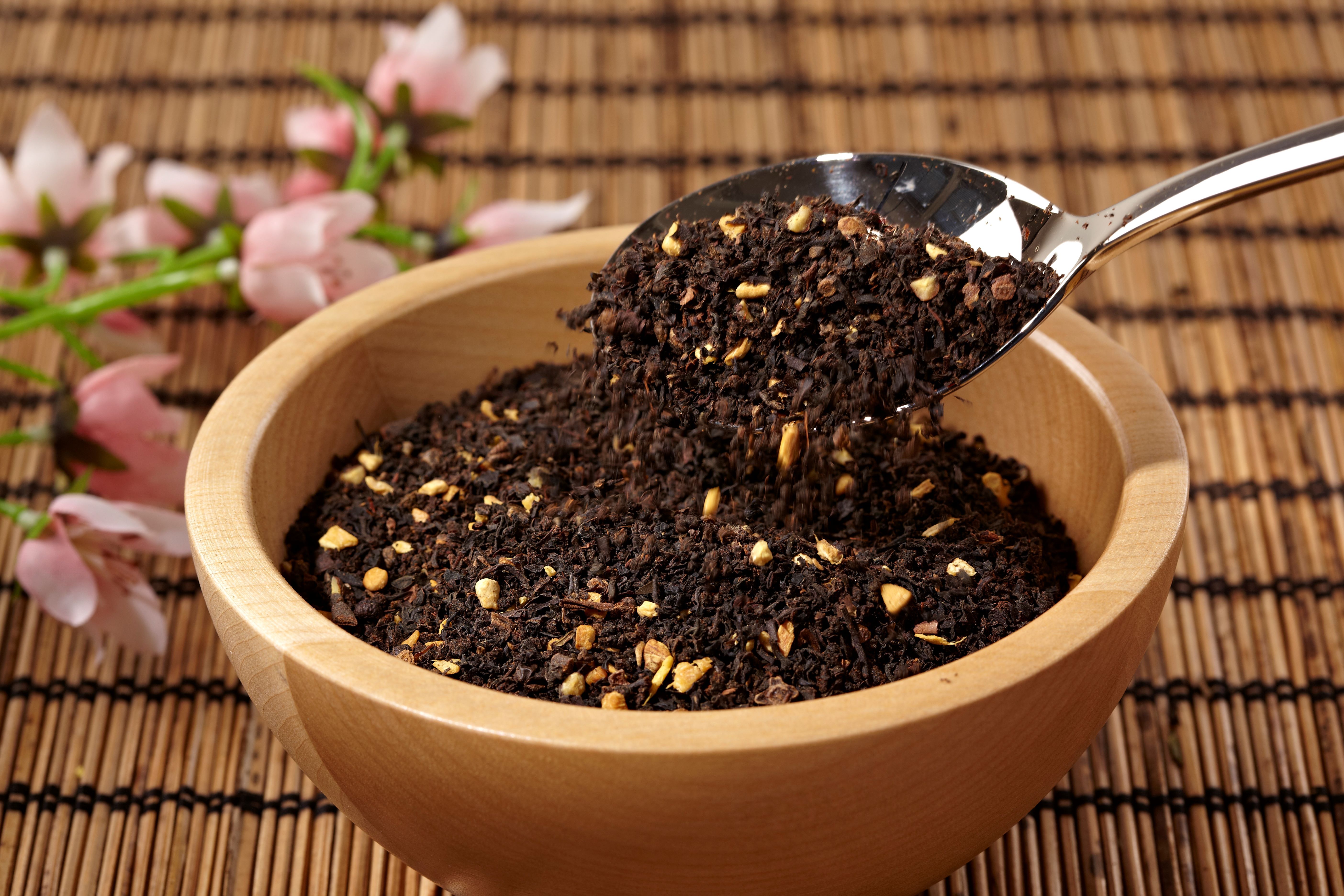 Holzschale mit losem Tee. Ein Löffel hebt Tee aus der Schale. Blüten im Hintergrund.