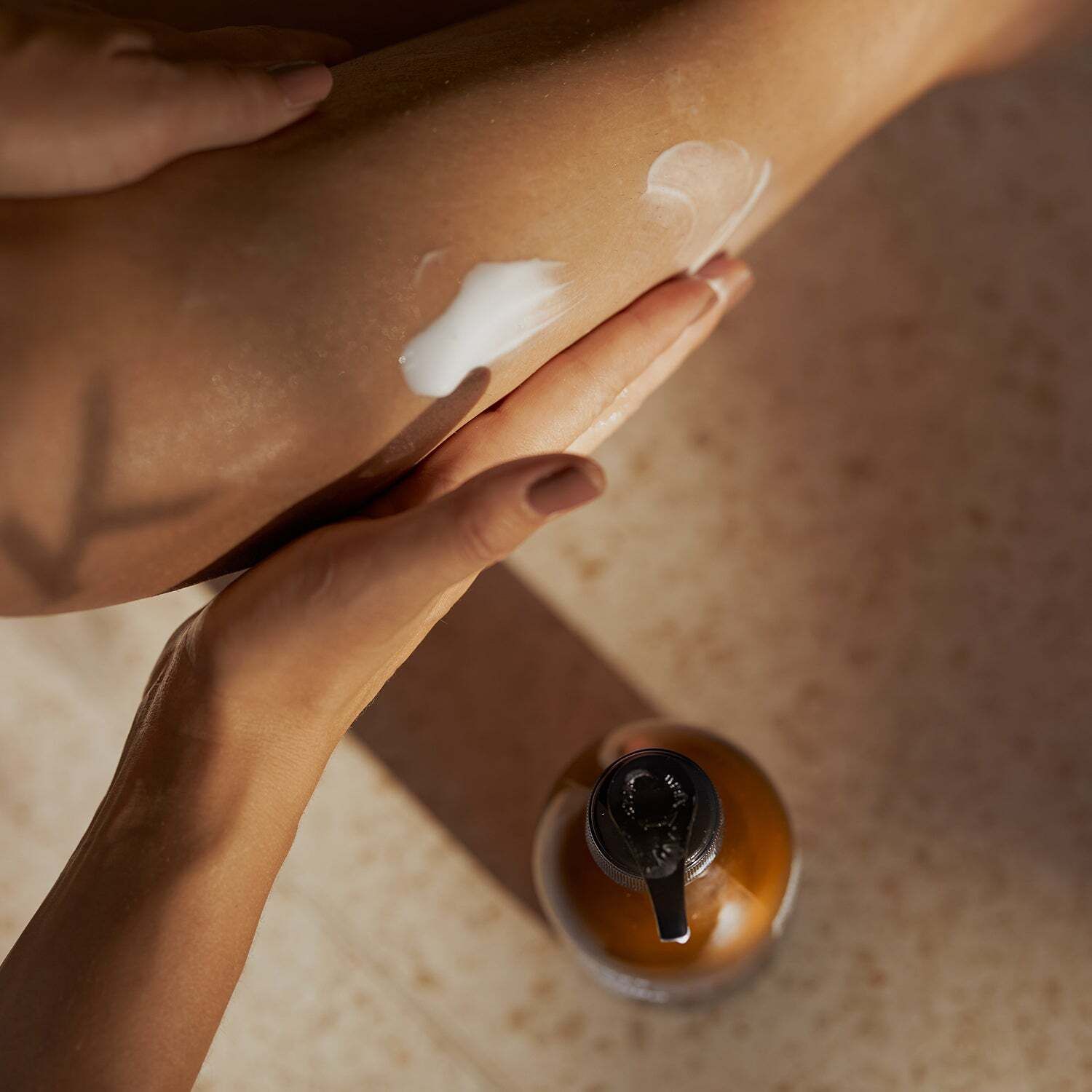 Ansicht von oben auf eine braune Flasche mit schwarzem Pumpkopf und eine Hand, die Lotion auf einen Arm aufträgt.
