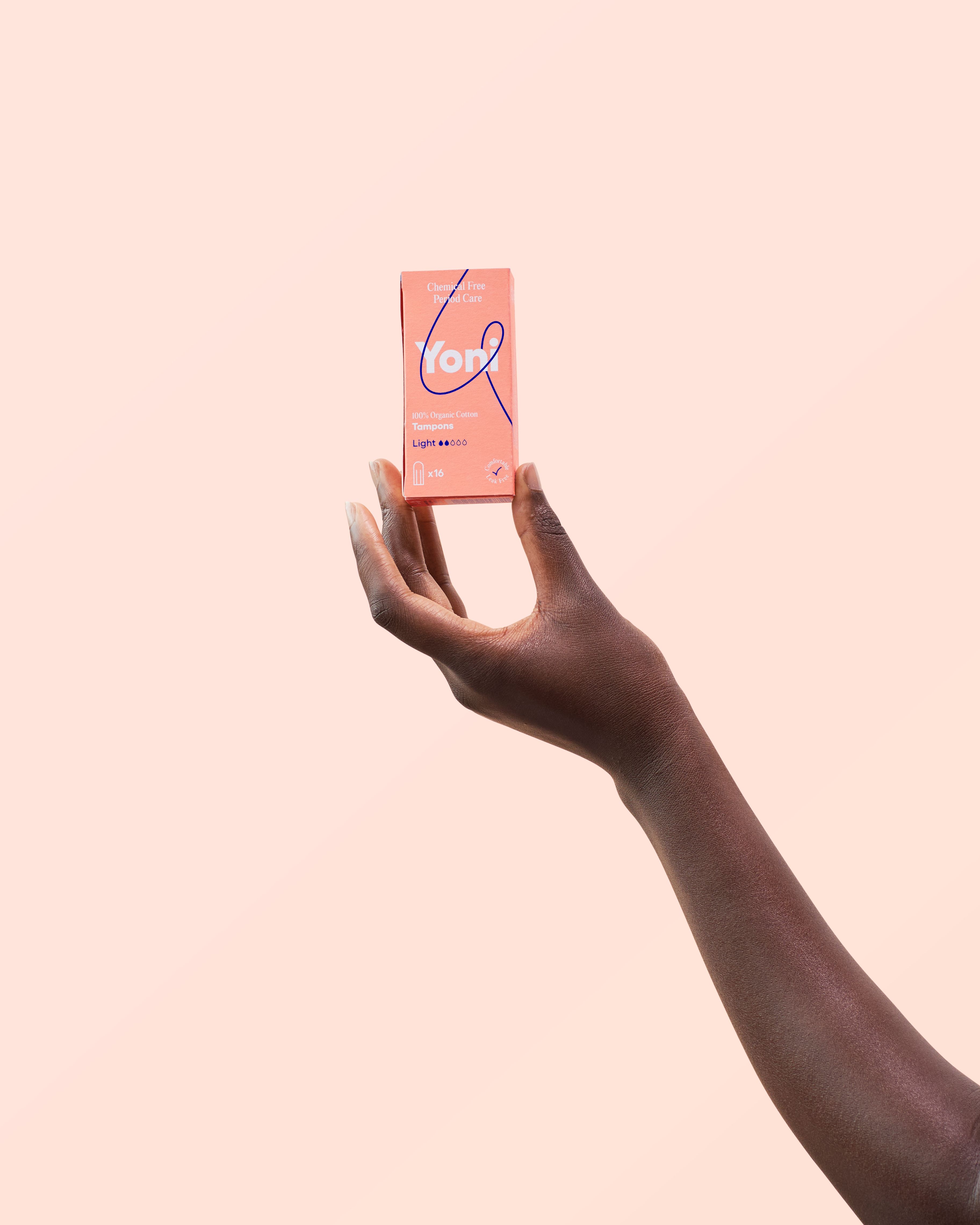 Hand hält eine Packung Tampons. Aufdruck: Yoni, 100% Bio-Baumwolle, Light. Hintergrund: hellrosa.