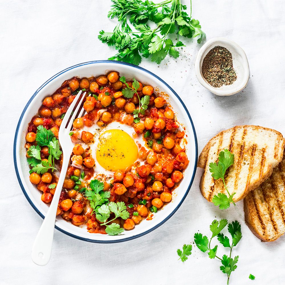 Teller mit Kichererbsen, Tomatensauce und Ei. Dazu Brot und Kräuter. Gewürze in Schale.