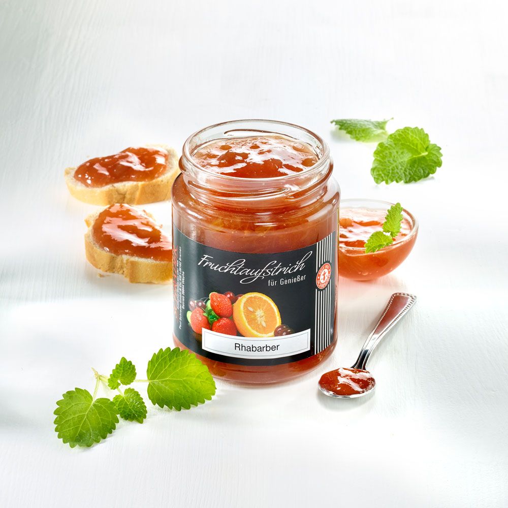 Glas mit Schrader Fruchtaufstrich Rhabarber, daneben Brotscheiben, Löffel und Minze. Aufschrift: Fruchtaufstrich für Genießer.