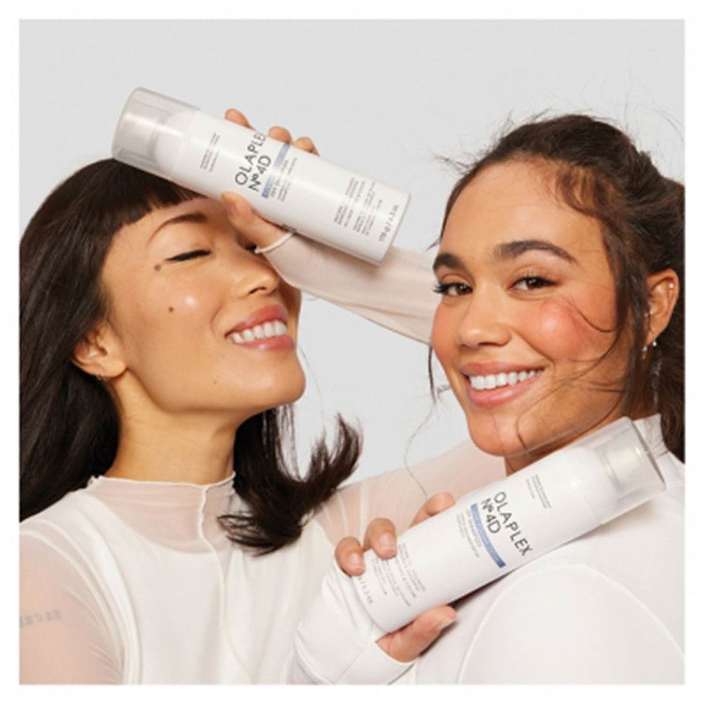 Zwei Frauen halten Olaplex Nº4D Trockenshampoo-Sprays. Sie lächeln.