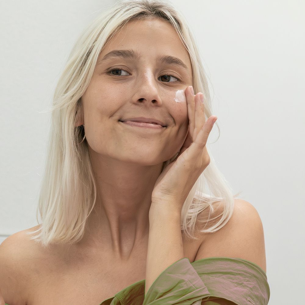 Junge Frau mit blonden Haaren trägt Balsam auf die Wange auf. Sie lächelt. Helle Umgebung.
