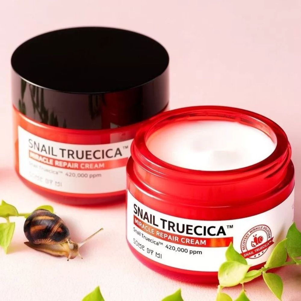 Zwei SOMEBYMI Snail Truecica Miracle Repair Cream Dosen, eine geöffnet, daneben eine Schnecke und Blätter.