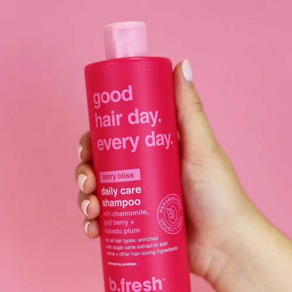 Rosa Shampoo-Flasche in einer Hand gehalten. Text: Good Hair Day. Every Day. Berry Bliss Daily Care Shampoo. Marke: b.fresh. Paraben Free, Cruelty Free.