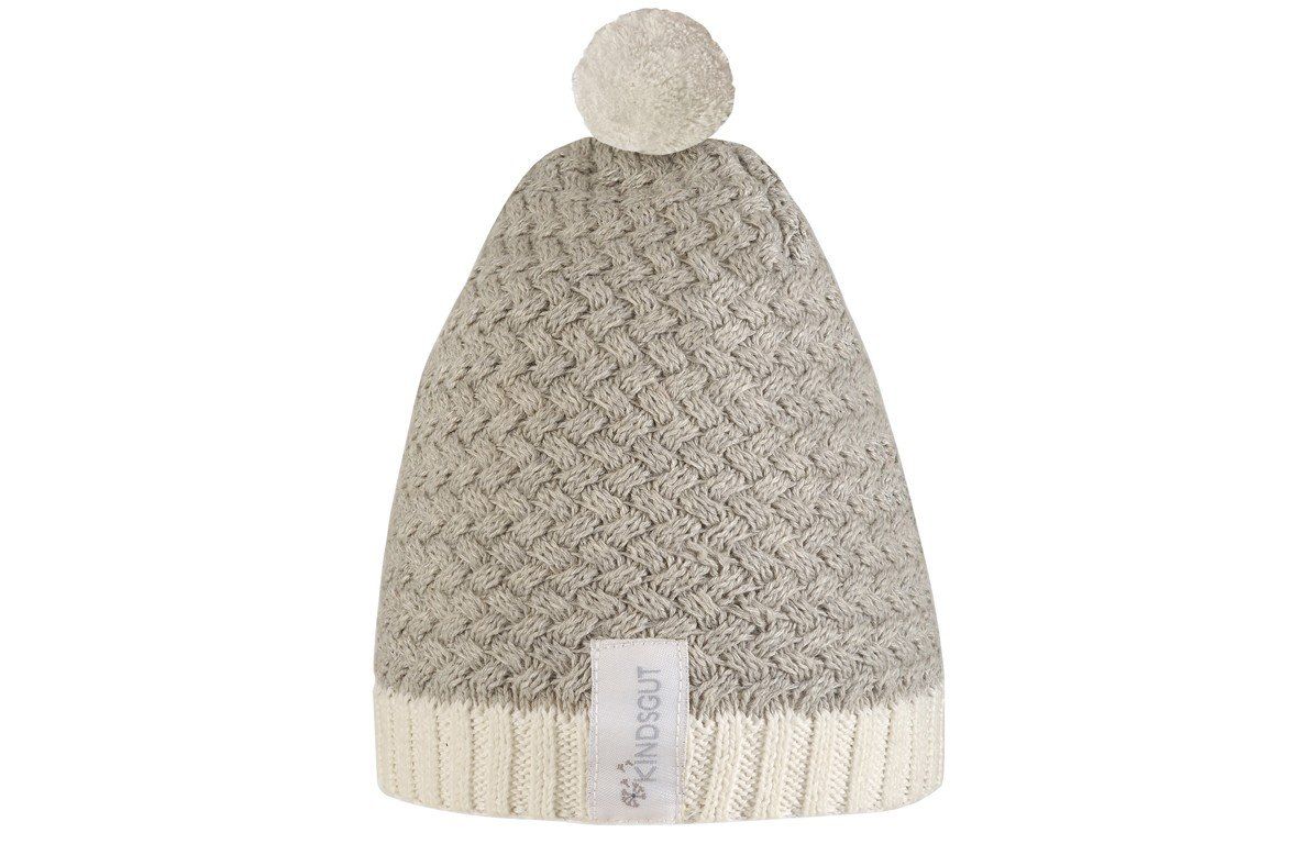 Grau-weiße Bommelmütze mit Label. Strickmuster, Bommel oben. Label mit Kindsgut-Logo.