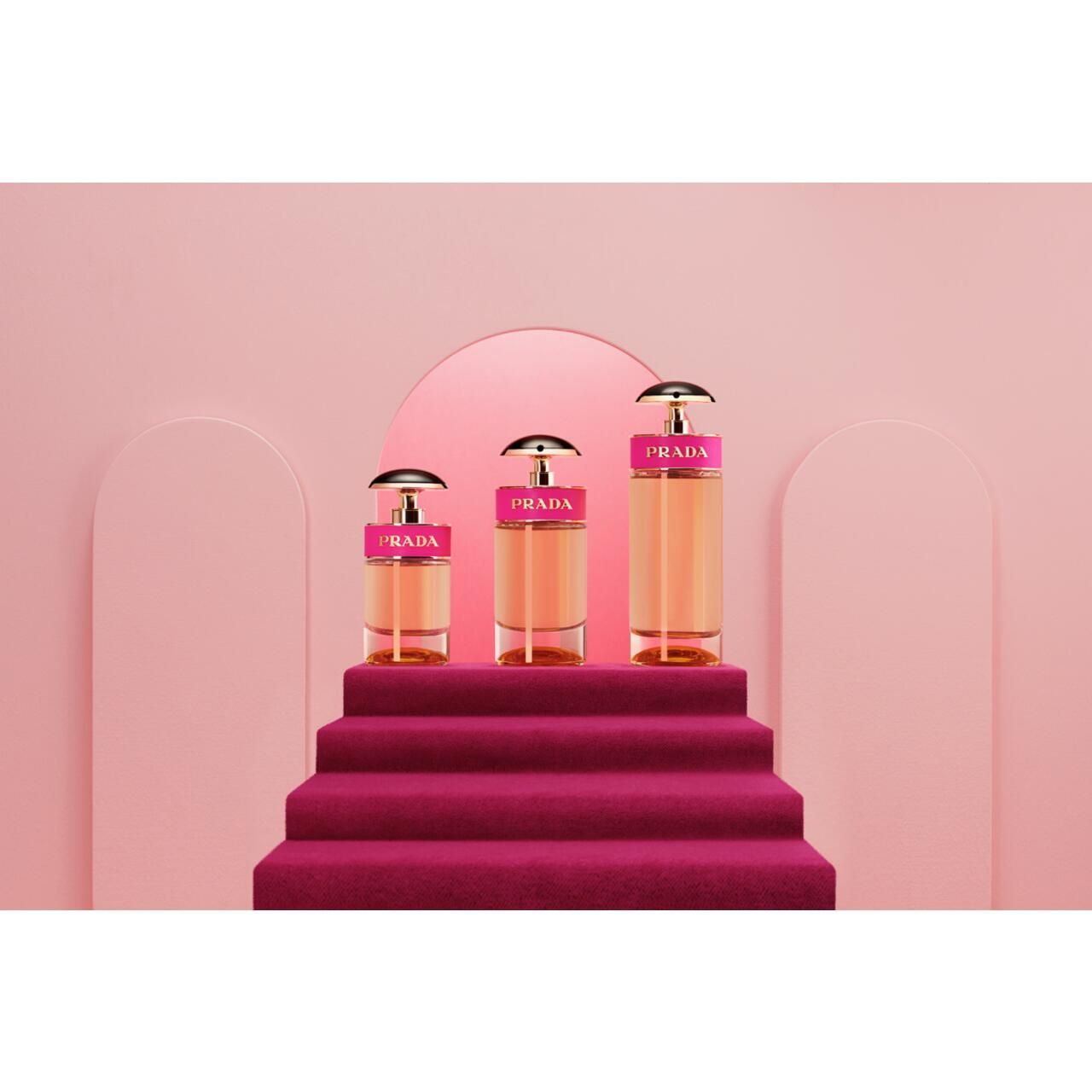 Drei Parfümflakons auf einer Treppe. Pinkfarbener Hintergrund. Flakons mit goldfarbenen Elementen und pinkfarbenem Band.