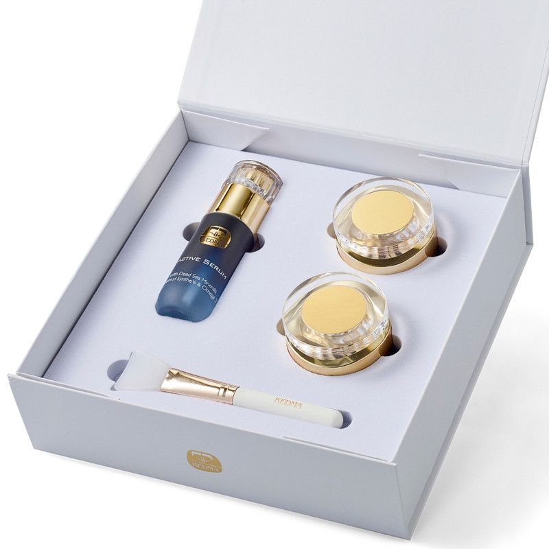 Geschenkbox mit drei Kosmetikprodukten: Serum, zwei Cremes und ein Pinsel. Die Produkte sind in einer weißen Box angeordnet.