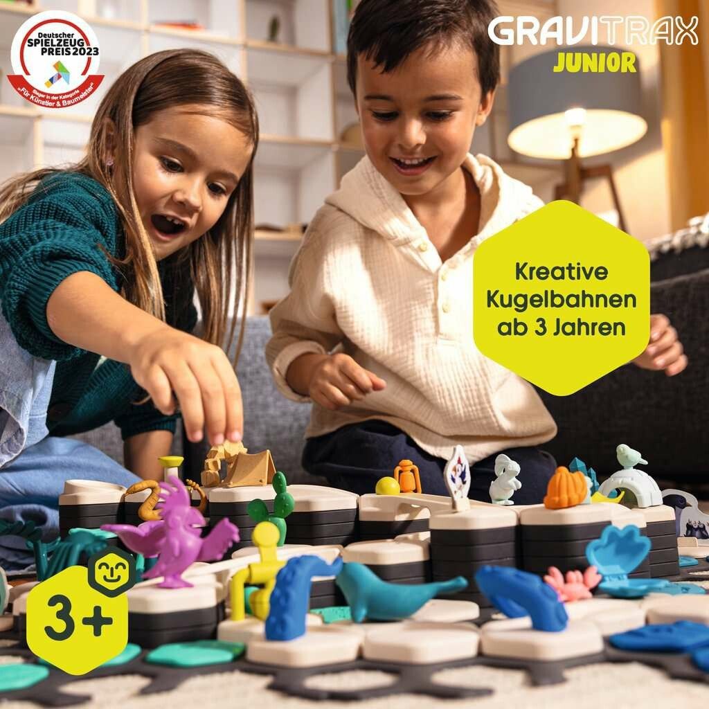 Due bambini giocano con il Ravensburger GraviTrax Junior Starter-Set XXL Planet. Il gioco è allestito sul pavimento, con vari elementi e personaggi. Il gioco è adatto a bambini dai 3 anni in su.