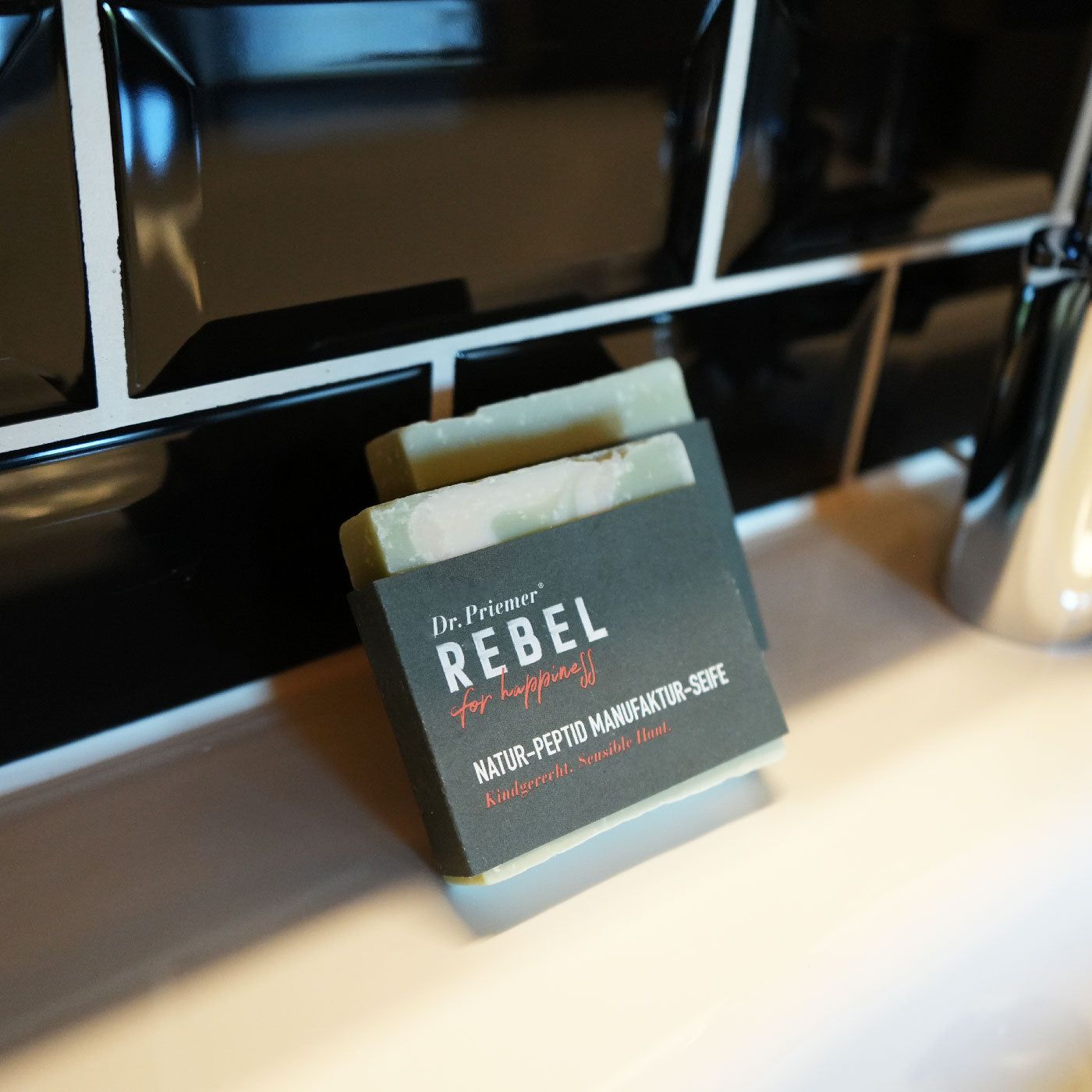 Zwei Seifen auf einer weißen Oberfläche, vor schwarzen Fliesen. Aufschrift: Dr. Priemer, REBEL for happiness, Natur-Peptid Manufaktur-Seife.