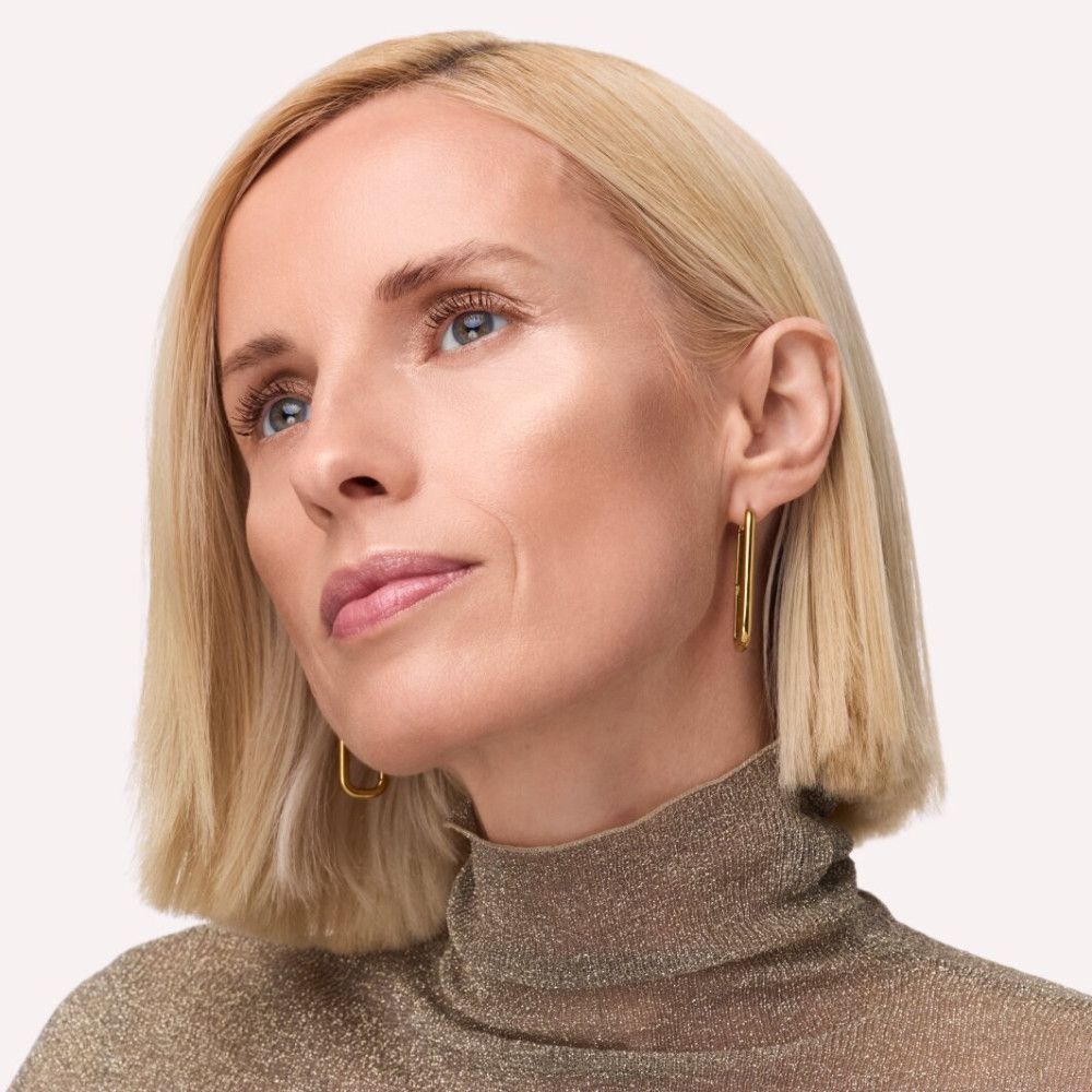 Porträt einer Frau mit Highlighter aufgetragen. Die Haut hat einen Glanz. Sie trägt goldene Ohrringe und einen Rollkragenpullover.