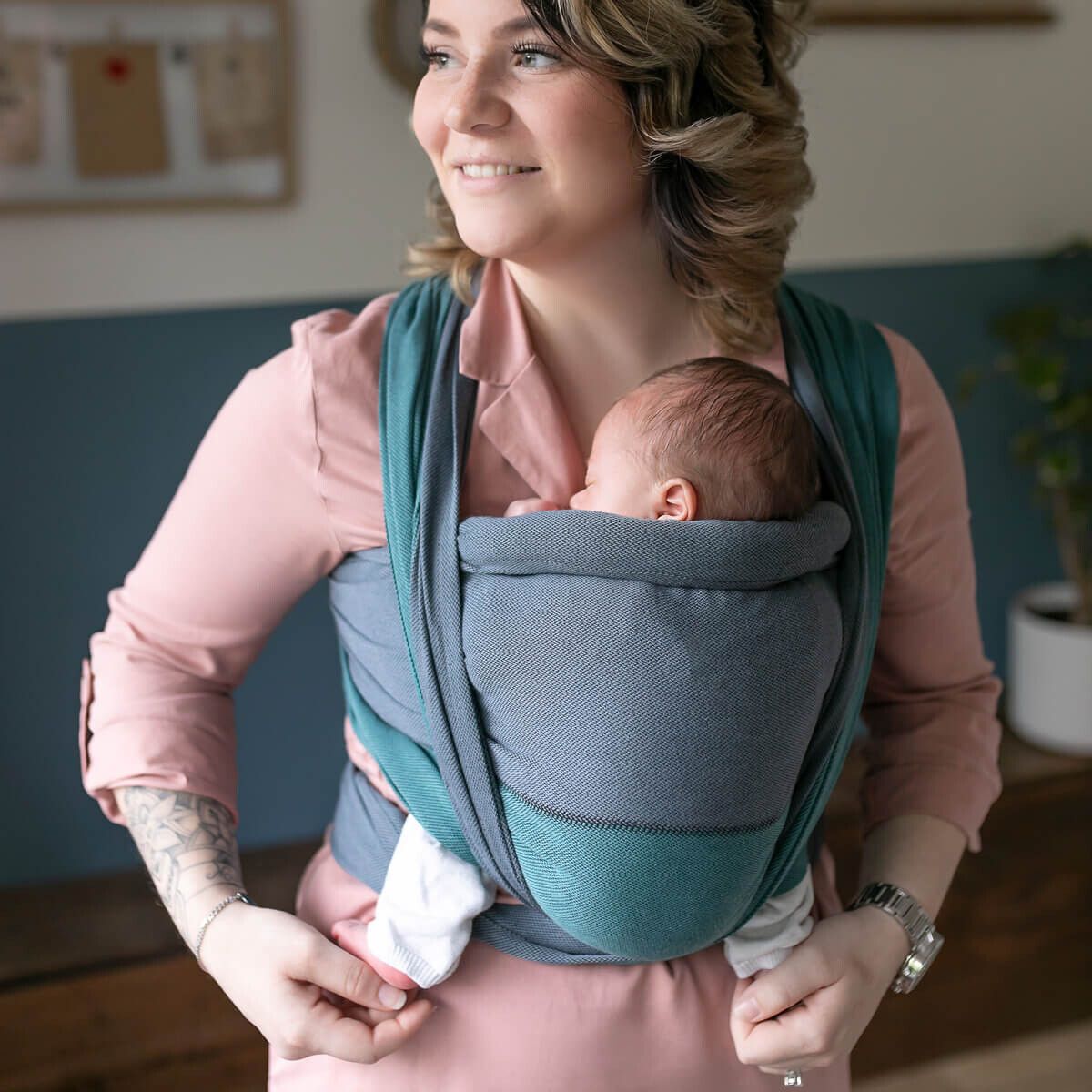 Frau hält Baby in blau-grüner Trage. Baby schläft. Frau trägt rosa Hemd. Innenraum.