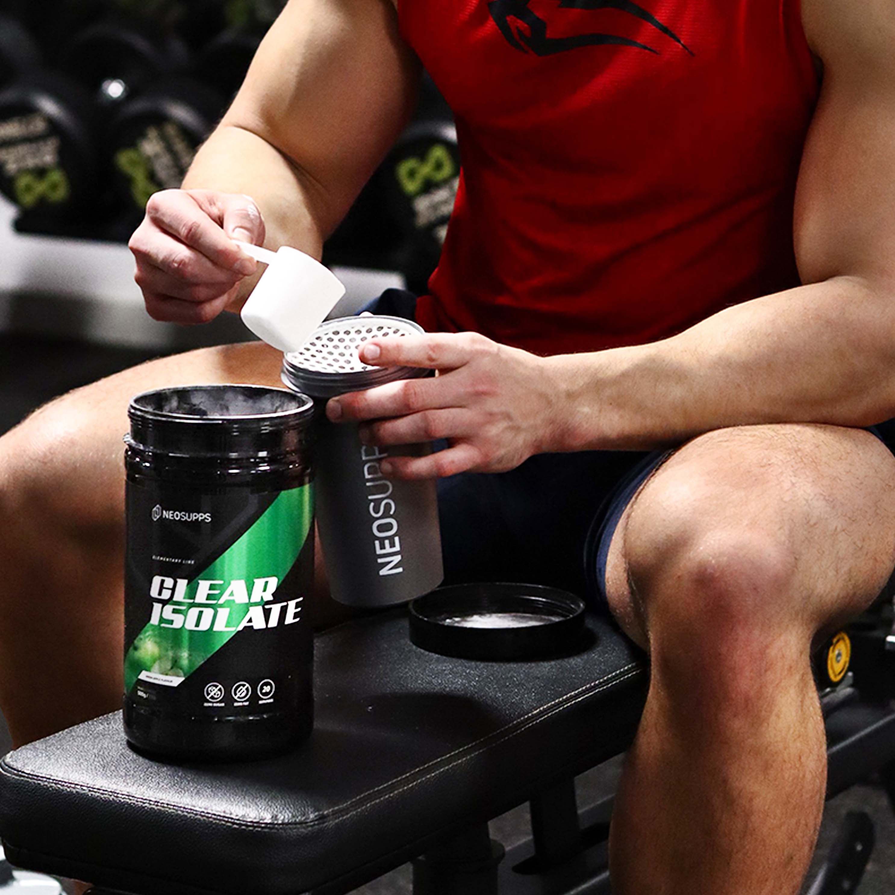 Mann öffnet Dose mit grünem Etikett. Aufschrift CLEAR ISOLATE und NEOSUPPS. Shaker und Dose auf Bank in Fitnessstudio.