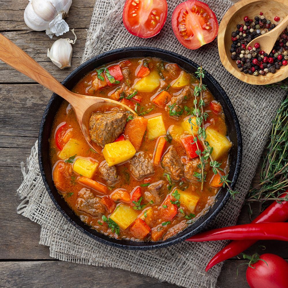 Gulasch in schwarzer Pfanne. Fleisch, Kartoffeln, Karotten, Paprika. Holz-Löffel. Holz-Schale mit Pfefferkörnern. Rote Chili.