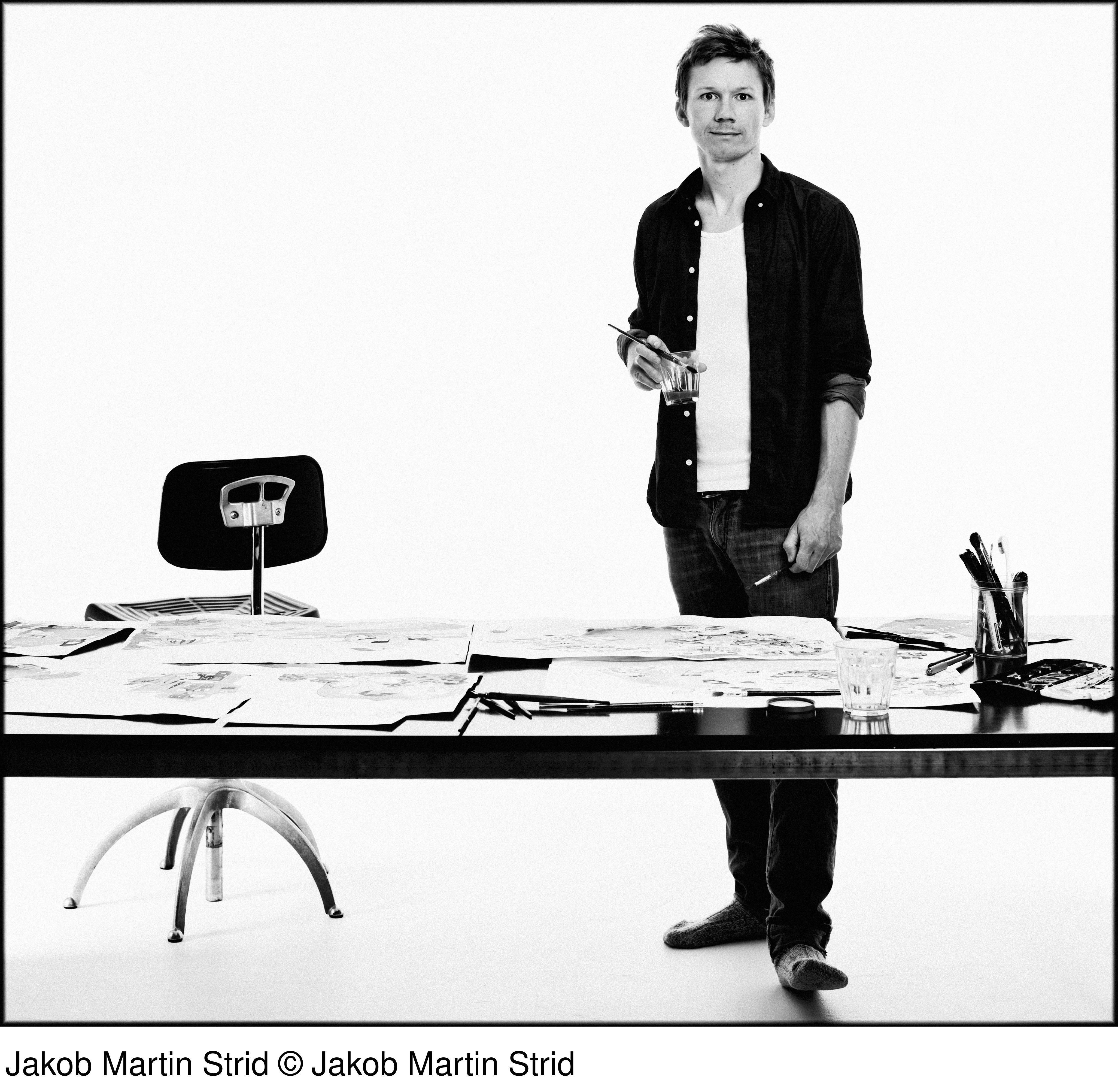 Schwarz-Weiß-Foto von Jakob Martin Strid. Er steht an einem Tisch mit Zeichnungen und hält einen Pinsel.