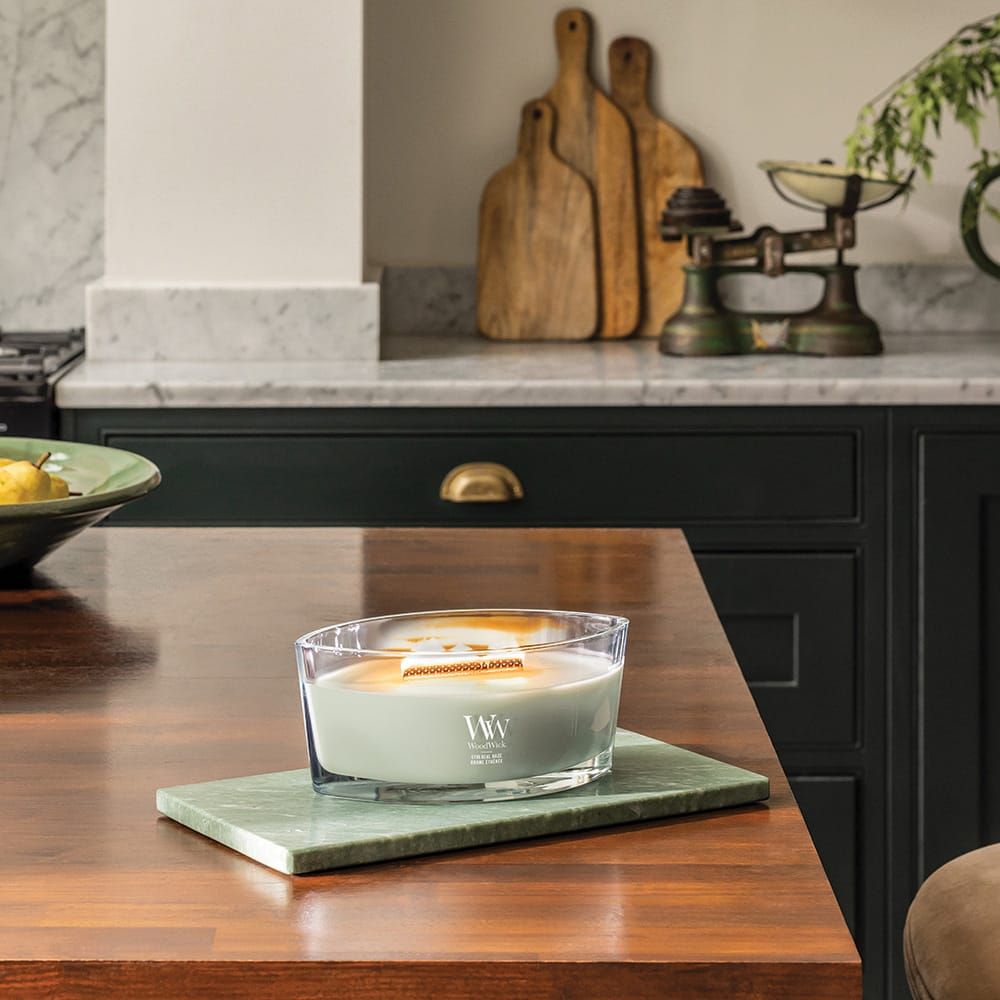 Ellipsenförmige Kerze auf grüner Platte. Glasbehälter mit hellgrünem Wachs. Marke WoodWick, Text: Ethereal Haze. Küche im Hintergrund.