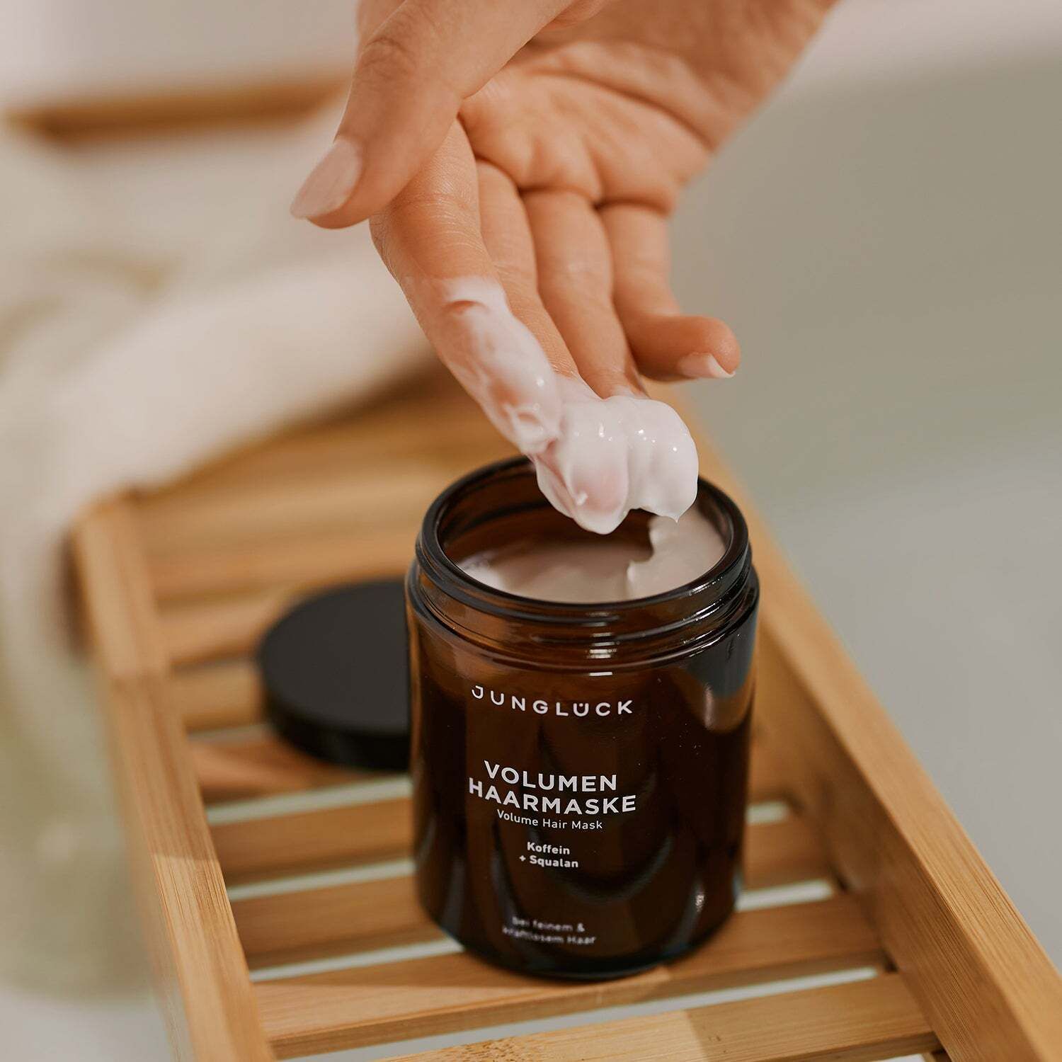 Hand entnimmt Haarmaske aus Dose. Dose: JUNGLÜCK Volumen Haarmaske. Auf Holztablett. Produkttext: Koffein + Squalan. Für feines & kraftloses Haar.