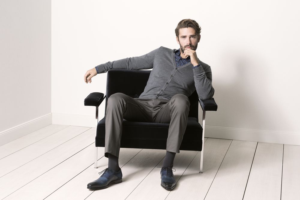 Mann sitzt in einem Stuhl. Er trägt graue Hose, Cardigan und blaue Schuhe. Hält Hand am Kinn.