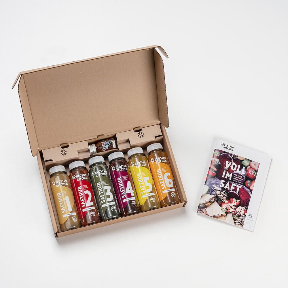 Geöffnete Verpackung mit 6 Flaschen Kloster Kitchen Saftkur und einem Shot. Daneben ein Buch mit dem Titel "Voll im Saft".