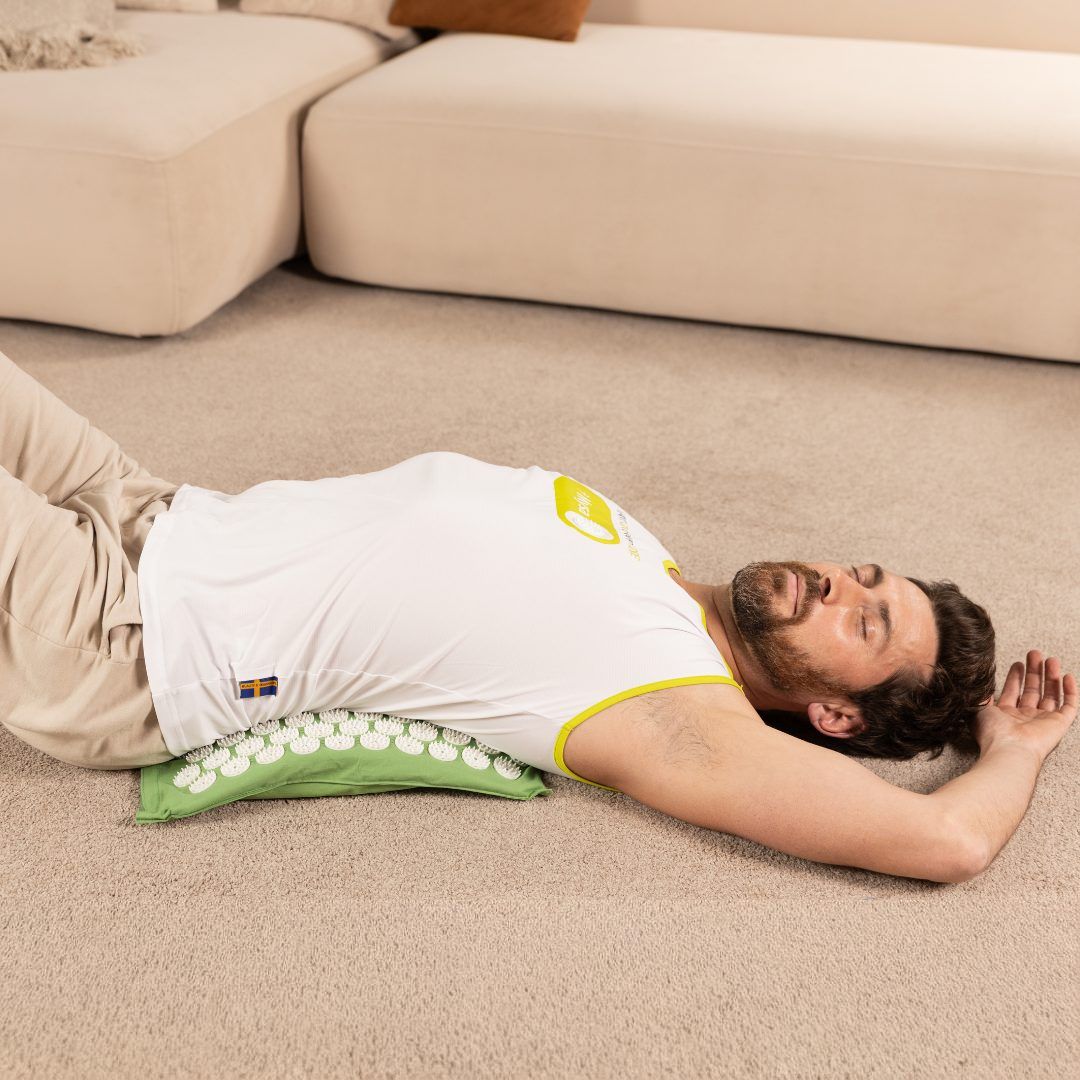 Mann liegt auf einer grünen Matte mit Noppen. Er trägt ein weißes T-Shirt mit gelben Akzenten.