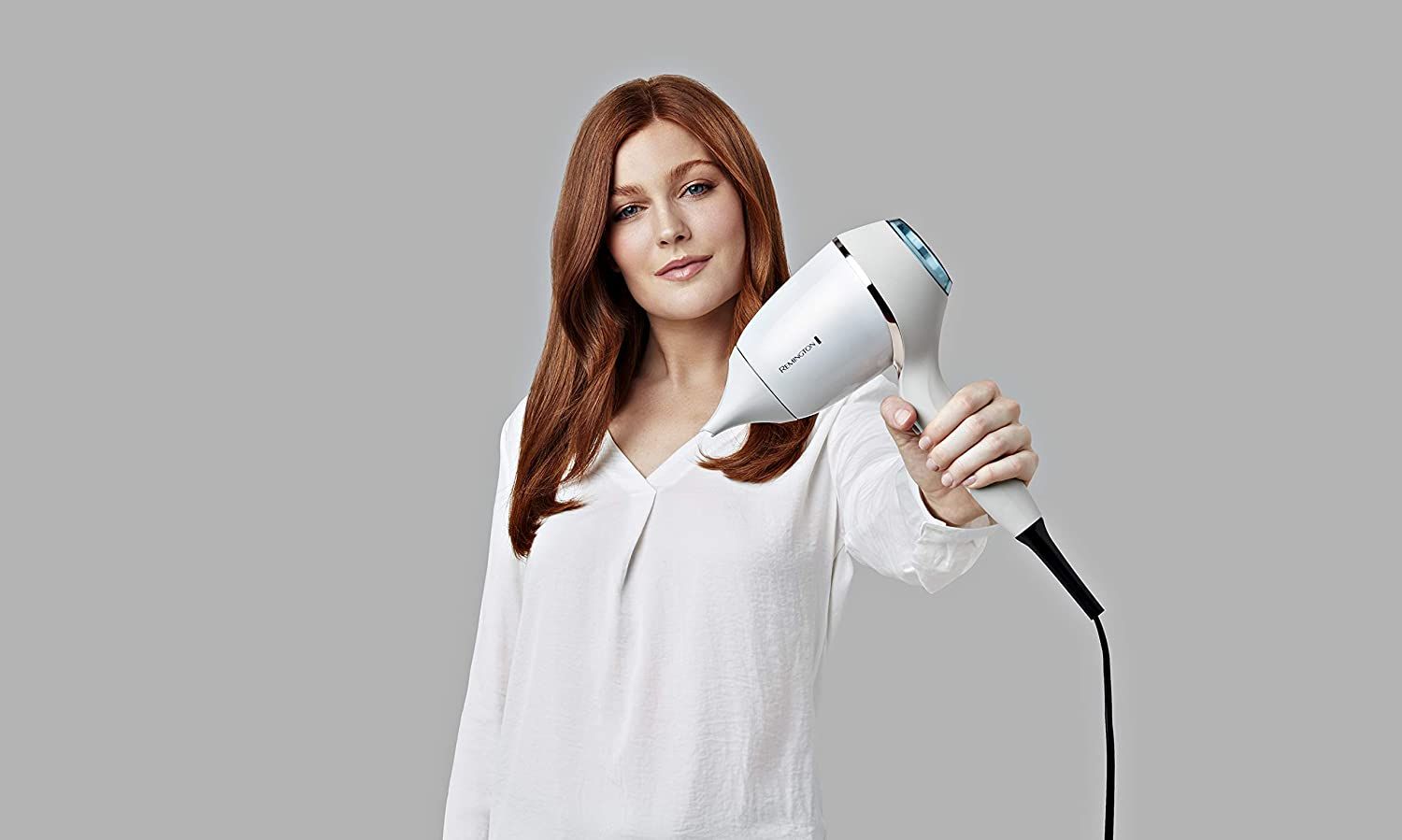 Frau hält weißen Haartrockner. Grauer Hintergrund. Sie trägt ein weißes Oberteil.