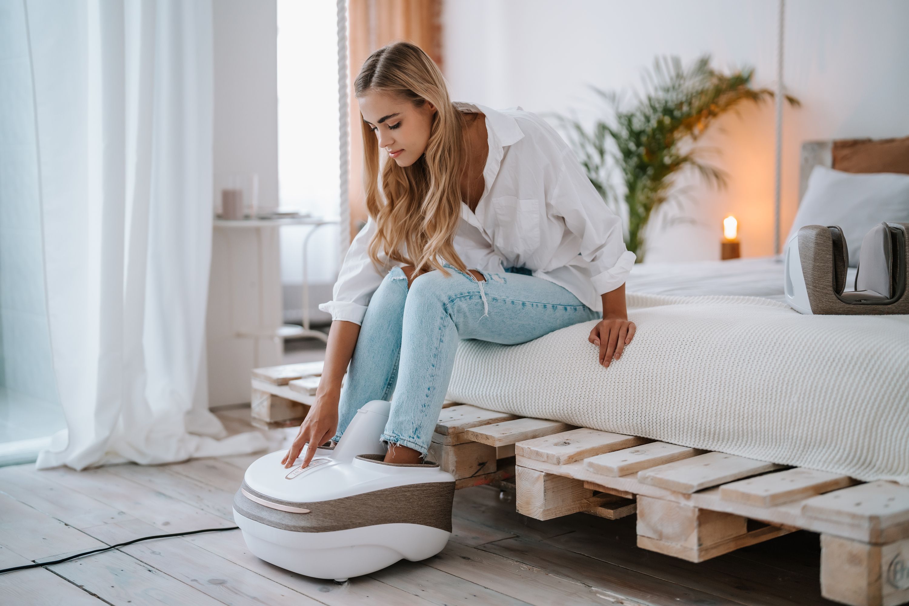Junge Frau sitzt auf einem Bett und bedient ein weißes Massagegerät. Das Gerät steht auf dem Boden.