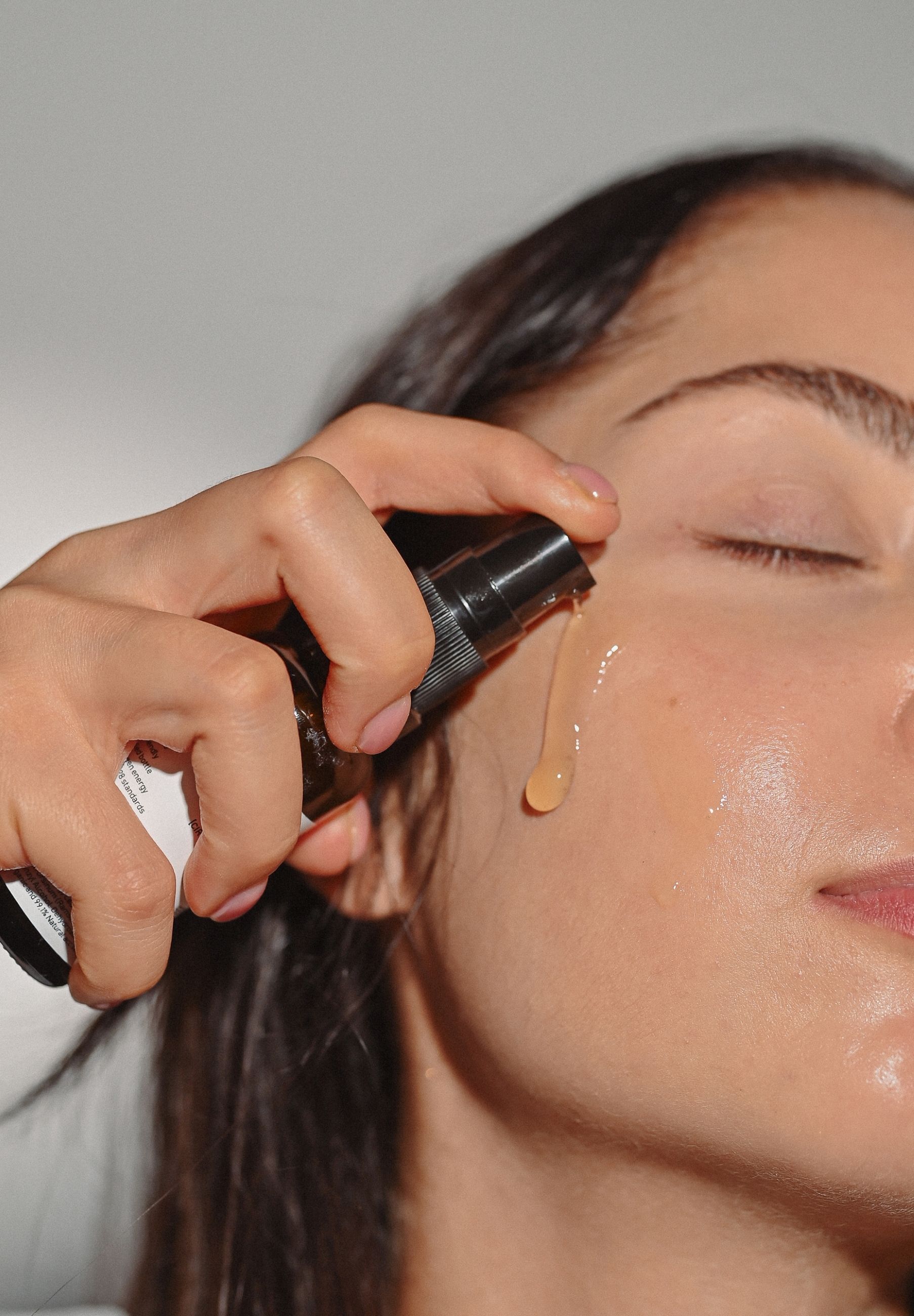 Person hält Flasche und sprüht Serum auf die Haut. Serum tropft auf die Wange. Haut mit Serum benetzt.