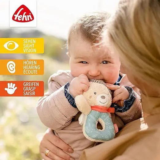 Baby hält Plüsch-Bärchen-Rassel im Mund. Fehn-Logo oben links. Visuelle, auditive und taktile Icons.