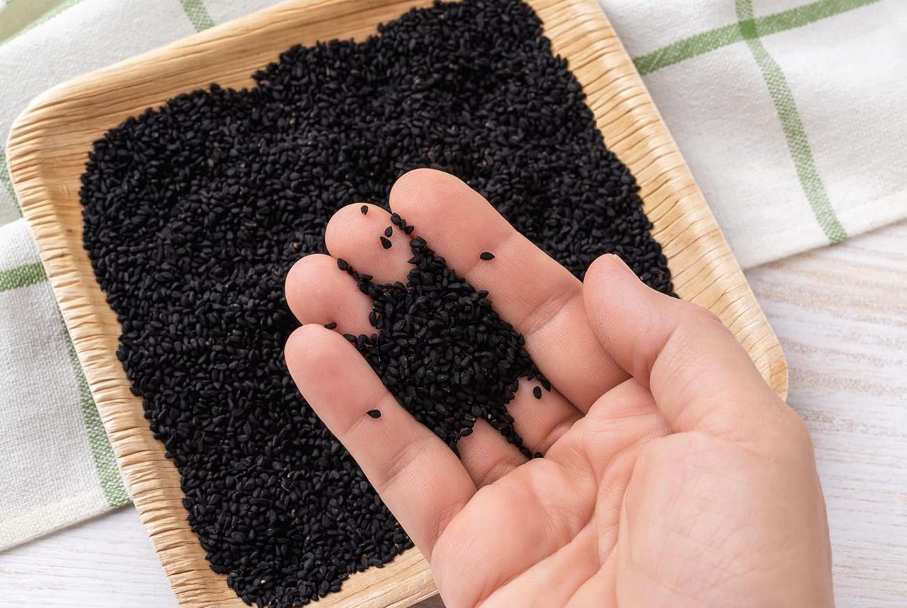 Hand mit schwarzen Samen auf einem Holztablett. Einige Samen auf den Fingern. Hintergrund: weißer Tisch, grünes Tuch.