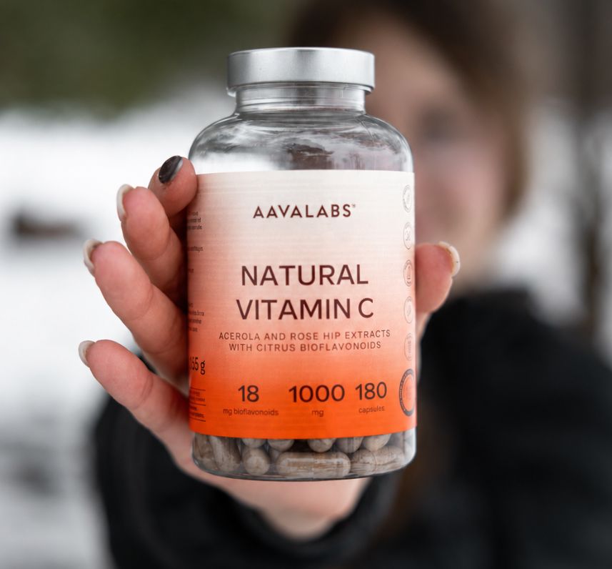 Hand hält eine Flasche Aavalabs natürliches Vitamin C 1000 mg vor verschwommenem Hintergrund.